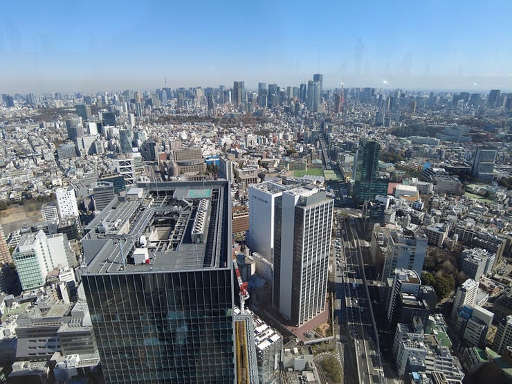 Aerial view of a cityscape featuring modern skyscrapers under a clear blue sky. Dense urban layout, visible streets, and rooftop structures. Tokyo Skytree & Tokyo Tower in the distance
