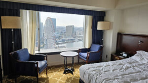Hotel room with a view of a Ferris wheel outside. Two blue armchairs, a small round table, and a rumpled bed, creating a cozy ambiance.