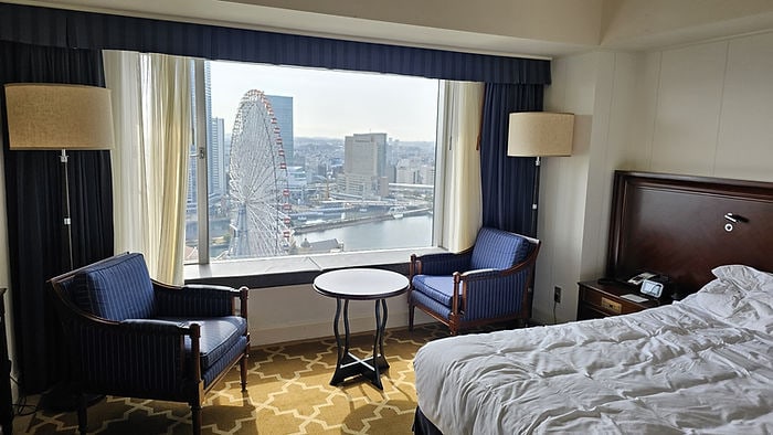 Hotel room with a view of a Ferris wheel outside. Two blue armchairs, a small round table, and a rumpled bed, creating a cozy ambiance.
