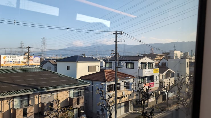 View from a train window shows houses, power lines, and mountains under a clear sky. A billboard in Japanese is visible. Calm suburban scene.