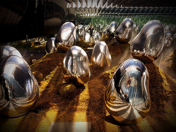 Shiny, smooth metallic sculptures on mossy mounds in a modern gallery, reflecting light and surroundings. The mood is serene and reflective.