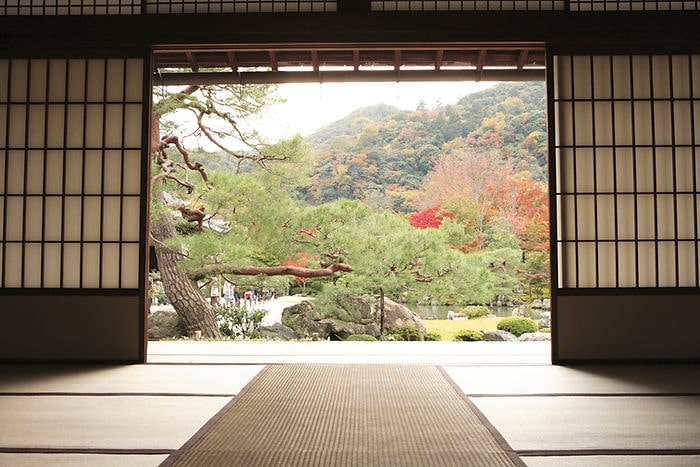 View from a traditional room with tatami mats. Open shoji doors reveal a tranquil garden with autumn trees and distant mountains.