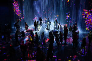 People explore an immersive art exhibit with colorful digital projections of flowers and abstract patterns on walls and floors, creating a surreal atmosphere.