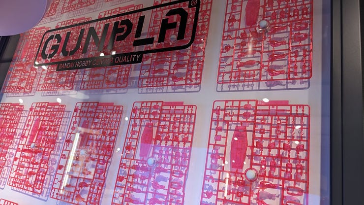 Red model kit sprues in a display case with the text "GUNPLA" and "Bandai Hobby Center Quality" visible. Bright, vibrant lighting.