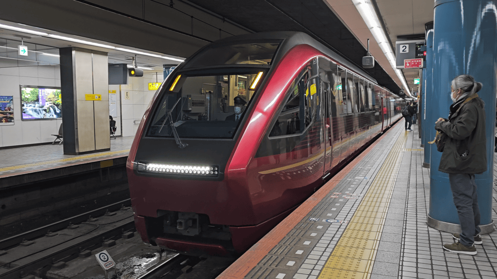 The sleek, metallic red Kintetsu 80000 series Hinatori luxury train traveling along scenic rail tracks through the Japanese countryside.
